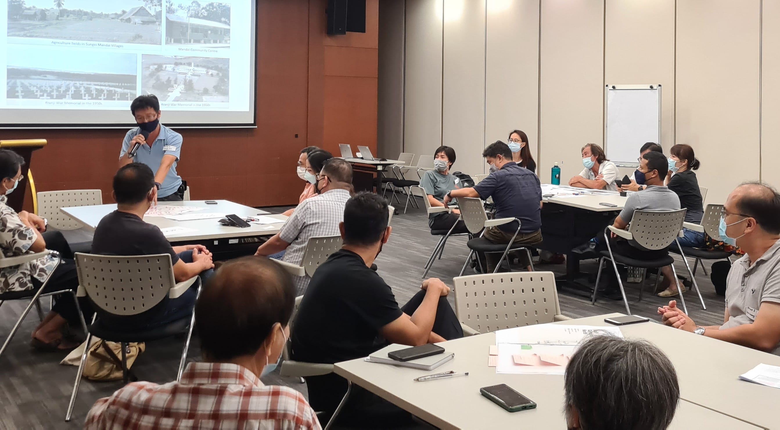 Several people sit around tables in a conference room, listening attentively to a presenter standing by a screen.